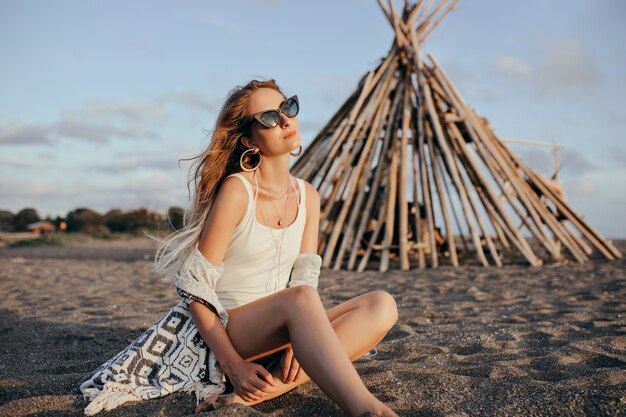 Elegante Frau in der schwarzen Sonnenbrille, die am Strand sitzt und Himmel betrachtet.