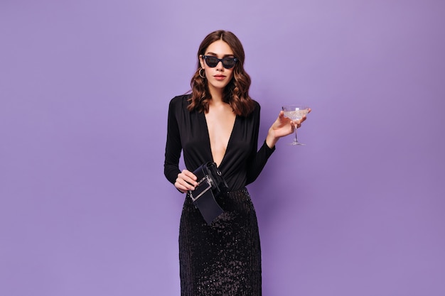 Elegante frau in sonnenbrille und schwarzem kleid hält martini-glas