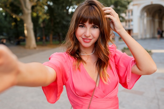 Elegante Frau mit den perfekten welligen Haaren, die Selbstporträt mit der Kamera machen. Rosa Kleid tragen.