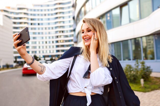 Elegante Geschäftsfrau, die auf der Straße in der Nähe des Büros posiert und einen trendigen, stilvollen Anzug und eine Ledertasche mit blonden Haaren trägt. Selfie machen und lächeln, per Video-Chat sprechen.