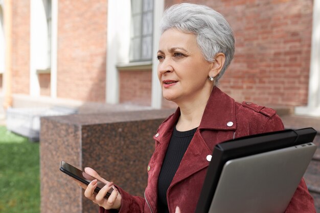 Elegante grauhaarige Frau mittleren Alters, die Laptop trägt, zum Geschäftstreffen geht und sich beeilt