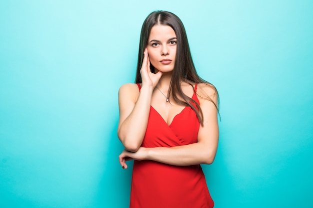 Elegante junge attraktive Frau, die rotes Sommerkleid mit den Händen am Kinn lokalisiert über pastellblauer Wand trägt.