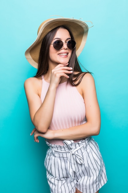 Elegante junge Frau, die Sommerkleid, Strohhut und Sonnenbrille trägt und an ihre Sommerferien denkt. Seitenansicht der Frau mit der Hand am Kinn, lokalisiert über pastellblauer Wand.