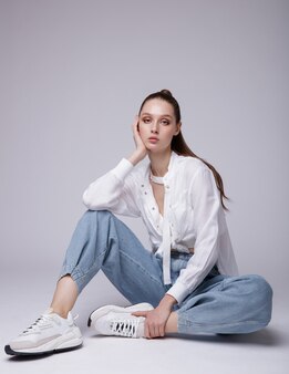 Elegante junge frau im weißen hemd turnschuhe blaue denim-jeans im hintergrund das modell sitzt