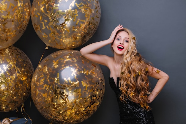 Elegante junge Frau mit langen blonden Haaren, die in der selbstbewussten Pose auf Neujahrsparty stehen. Innenporträt des charmanten Geburtstagskindes, das mit funkelnden Luftballons aufwirft.