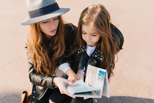 Elegante junge Frau und ihre Tochter verirren sich in einer fremden Stadt und schauen auf die Karte. Nettes kleines Mädchen mit ernstem Gesichtsausdruck bildet die Reiseroute mit schöner Mutter.