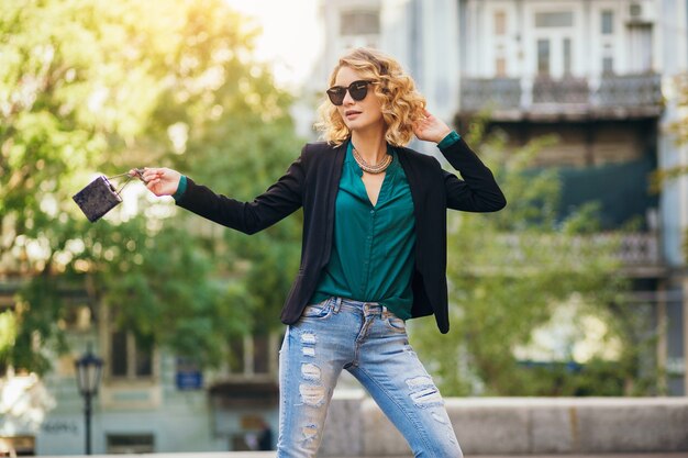 Elegante schöne Frau, die Sonnenbrille, Jacke, Jeans, grüne Bluse, Frühlingsmode-Trend trägt und kleine Tasche hält,