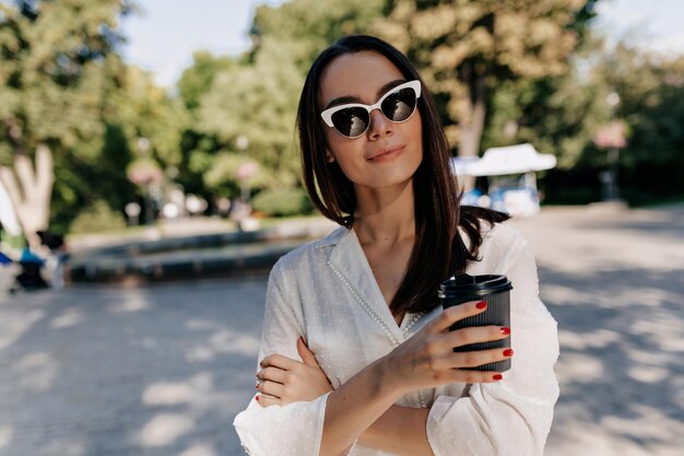 Elegante, stilvolle Frau mit glattem dunklem Haar, Sonnenbrille und weißem Hemd hält Tasse mit Kaffee und posiert im Sonnenlicht im grünen Sommerpark in der Stadt in die Kamera