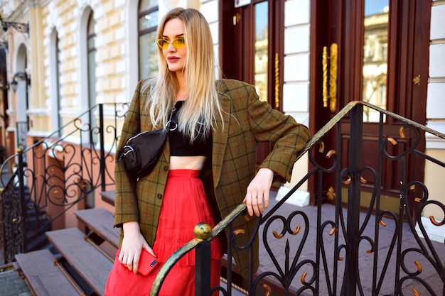 Elegante trendige blonde Frau, die auf der Straße nahe schönem altem Gebäude aufwirft, modisches trendiges Hipster-Outfit und Sonnenbrille tragend, Frühlingsherbststil.