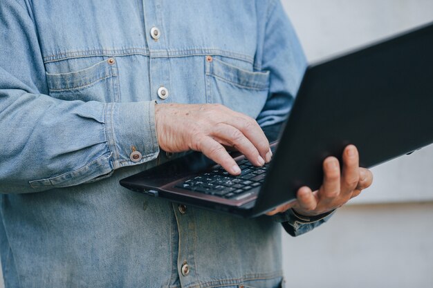 Eleganter alter Mann, der auf grauem Hintergrund steht und einen Laptop benutzt
