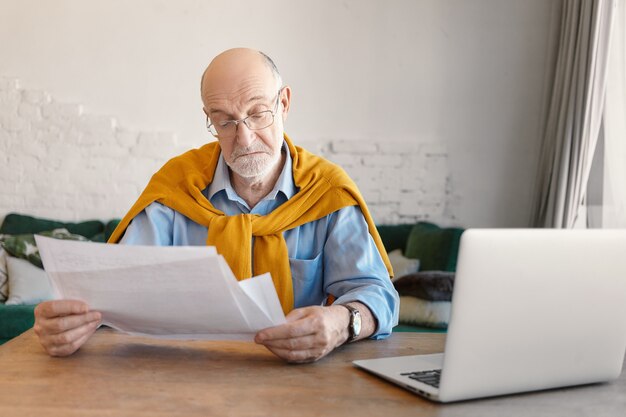 Eleganter bärtiger älterer Mann in rechteckigen Gläsern, die Papierblätter in seinen Händen studieren, häusliche Finanzen online zu Hause berechnen, unter Verwendung des elektronischen tragbaren Geräts im Wohnzimmerinnenraum