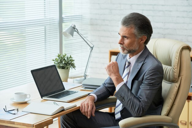 Eleganter Geschäftsmann am Bürotisch, der durchdacht dem Fenster betrachtet