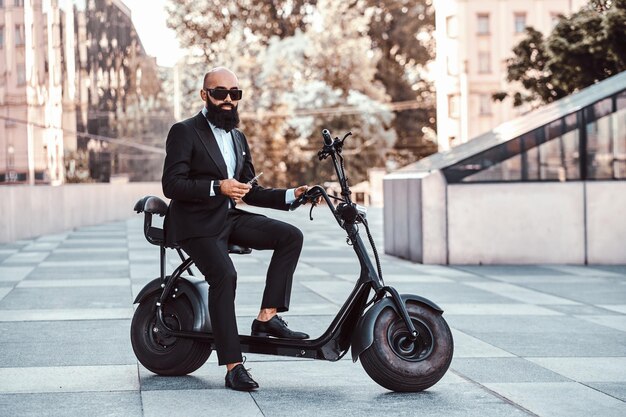 Eleganter glatzköpfiger Geschäftsmann mit Sonnenbrille raucht Verdampfer, während er auf seinem Elektrofahrrad sitzt.