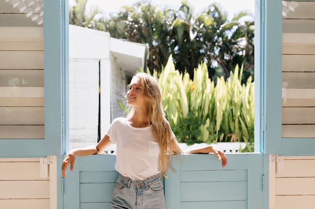 Elegantes blondes Mädchen, das mit geschlossenen Augen und inspiriertem Lächeln aufwirft. Foto der schlanken schönen Frau mit langen Haaren.