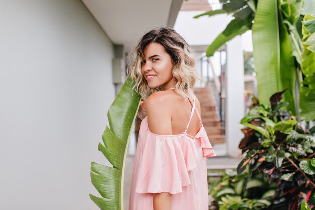 Elegantes gebräuntes Mädchen mit kurzer Frisur, die über Schulter schaut. Außenfoto der gutaussehenden kaukasischen jungen Frau in den rosa Kleidern, die im Hof aufwerfen.