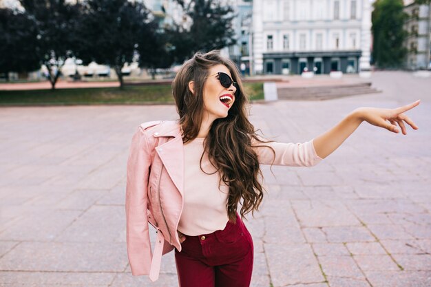 Elegantes Mädchen mit langer Frisur in weinigen Hosen, die Spaß in der Stadt haben. Sie hat eine rosa Jacke auf der Schulter und zeigt sich zur Seite.