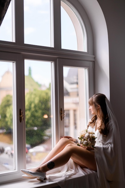 Elegantes Mädchen, während Robe und Schleier auf der Fensterbank sitzen