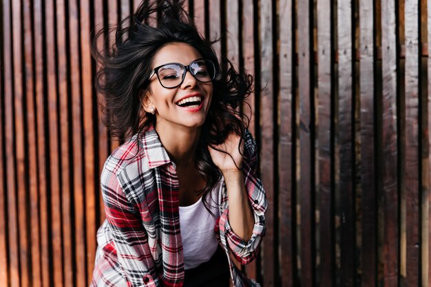 Elegantes schwarzhaariges Mädchen, das auf Holzwand tanzt. Charmante lateinamerikanische Frau in Gläsern, die postivie Gefühle ausdrücken und lachen.