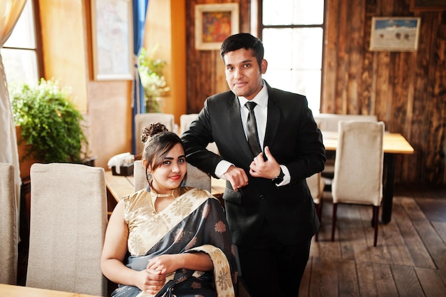 Elegantes und modisches indisches Freundespaar Frau im Saree und Mann im Anzug posierte im Innencafé