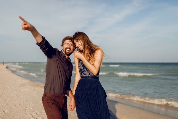 Elegantes verliebtes Paar, das am sonnigen Strand geht. Romantische Stimmung. Frau, die elegantes blaues Kleid mit Pailletten trägt. Ihr Mann zeigte auf etwas.