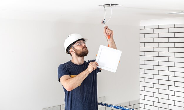 Elektriker Builder mit Bartarbeiter in einem weißen Helm bei der Arbeit, Installation von Lampen in der Höhe. Professionell in Overalls mit einem Bohrer auf der Reparaturstelle.