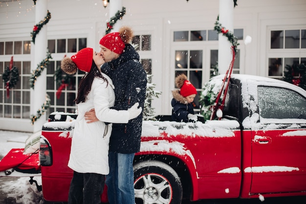Eltern in roten Hüten unter Schneefall im Freien küssen. Schönes Kind mit rotem Hut, das im Hintergrund im roten Pick-up spielt.