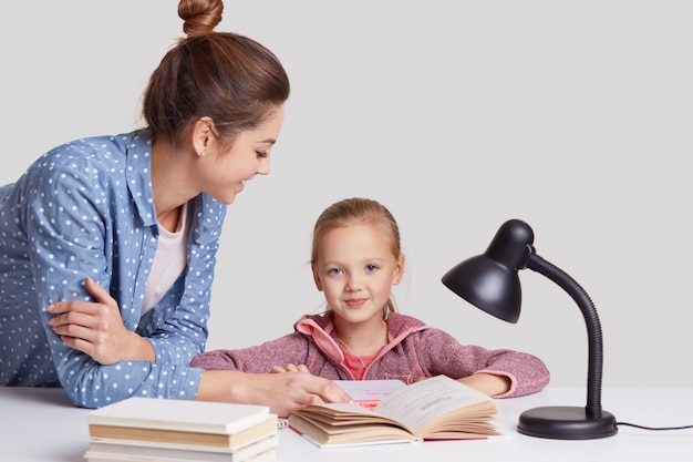 Elternschafts-, Studien- und Bildungskonzept, blauäugiges weibliches Kind sitzt am Arbeitsplatz, liest Buch zusammen mit Mutter, lernt Gedicht auswendig, posiert in gemütlichem Raum auf Weiß