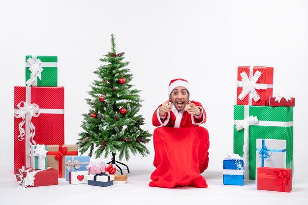 Emotional aufgeregter junger Mann verkleidet als Weihnachtsmann mit Geschenken und geschmücktem Weihnachtsbaum, der etwas auf weißem Hintergrund zeigt