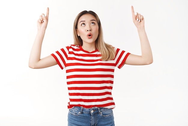 Emotional fasziniert, gutaussehend, blond, asiatisches mädchen, das die lippen faltet, sagen, wow, beeindruckt, nachdenklich, interessanter promo-anzeigenblick, der nach oben zeigt, hebt die zeigefinger nach oben, aufgeregter weißer hintergrund