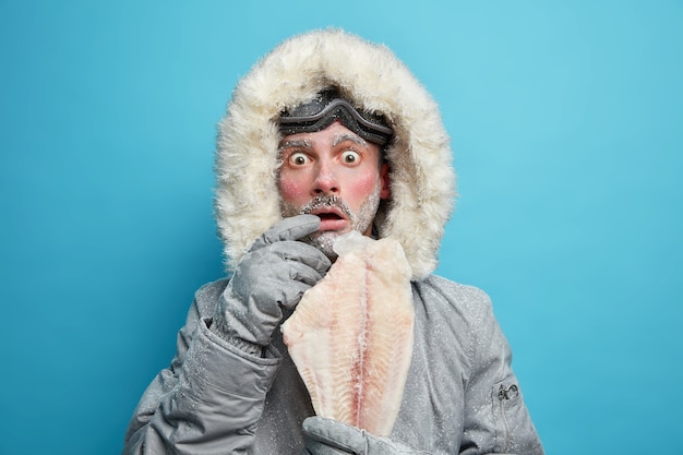 Kostenloses Foto emotional schockierter frostiger mann in winteroberbekleidung hält gefrorenen fisch fühlt sich bei niedrigen temperaturen im norden sehr kalt an.