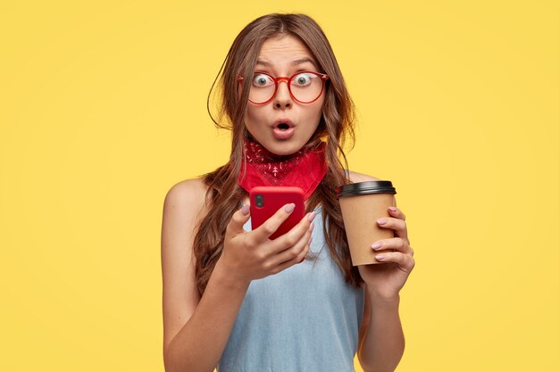 Emotional überrascht Teenager-Mädchen öffnet den Mund, fühlt sich erstaunt, hält Smartphone und Kaffee trinken, hat verblüfften Blick beeindruckt, erhält unerwartete Nachricht, isoliert über gelbe Wand