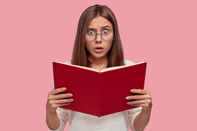 Emotional überraschte europäische Frau hält Lehrbuch, hat Angst Gesichtsausdruck, Sorgen vor dem Bestehen der Aufnahmeprüfung, trägt runde Brille über rosa Wand isoliert. Weibliches Mädchen mit rotem Buch