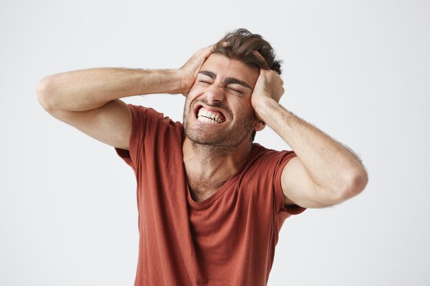 Emotional wütender muskulöser Mann, der rotes T-Shirt trägt, die Augen fest schließt und vor Schmerz oder vollem Unglauben schreit und Hände auf seinem Kopf hält. Negative menschliche Emotionen und Gefühle