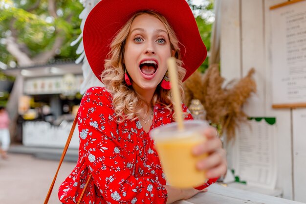 Emotionale attraktive stilvolle blonde lächelnde Frau in strohrotem Hut und Bluse Sommermode-Outfit, das natürlichen Fruchtcocktail-Smoothie trinkt