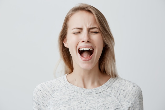 Emotionale blonde Frau öffnet ihren Mund und schreit laut, weil sie mit etwas unzufrieden ist, das Uneinigkeit und Ärger ausdrückt