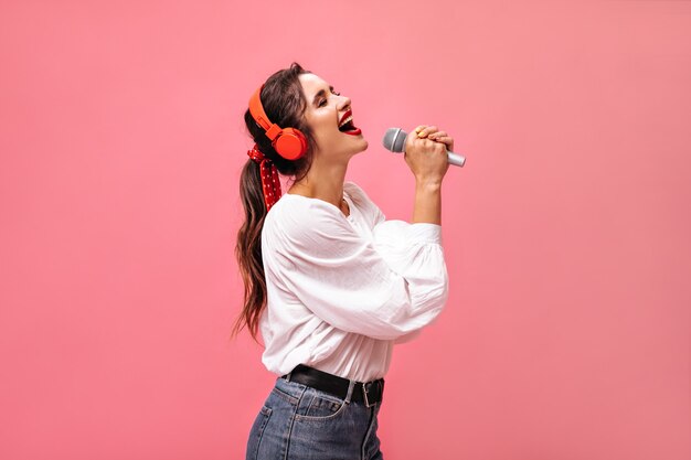 Emotionale Dame in den roten Kopfhörern, die im Mikrofon singen. Helles schönes Mädchen in der weißen Bluse und in den Jeans mit dem schwarzen Gürtel, der aufwirft.