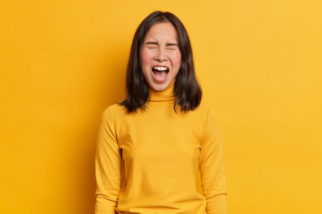 Emotionale dunkelhaarige asiatische Frau schreit laut drückt Wut hält Mund weit geöffnet trägt lässigen gelben Rollkragenpullover in einem Ton mit Studiohintergrund. Konzept menschlicher Emotionen und Gefühle