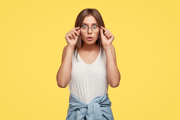 Emotionale entsetzte junge Brünette mit Brille, die gegen die gelbe Wand aufwirft