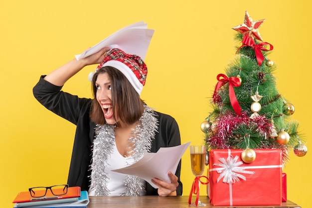 Emotionale erschöpfte Geschäftsdame im Anzug mit Weihnachtsmannhut und Neujahrsdekorationen, die Dokumente halten und an einem Tisch mit einem Weihnachtsbaum darauf im Büro sitzen