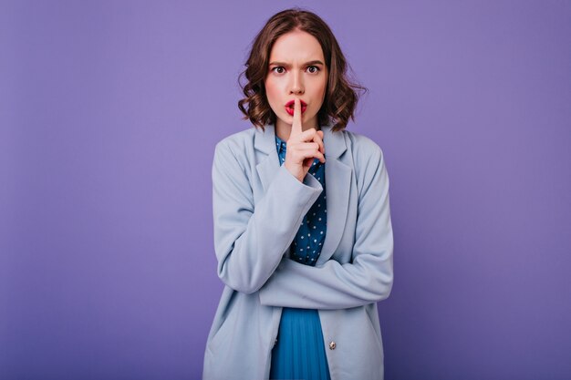 Emotionale Frau mit hellem Make-up, das Lippen mit Finger berührt. charmantes lockiges Mädchen im blauen Mantel, das auf lila Wand steht.