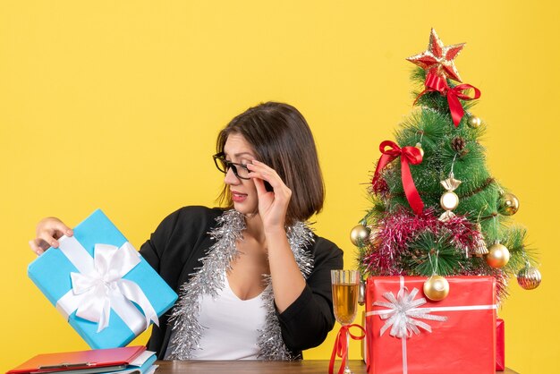 Emotionale Geschäftsdame im Anzug mit Brille, die ihr Geschenk betrachtet und an einem Tisch mit einem Weihnachtsbaum darauf im Büro sitzt