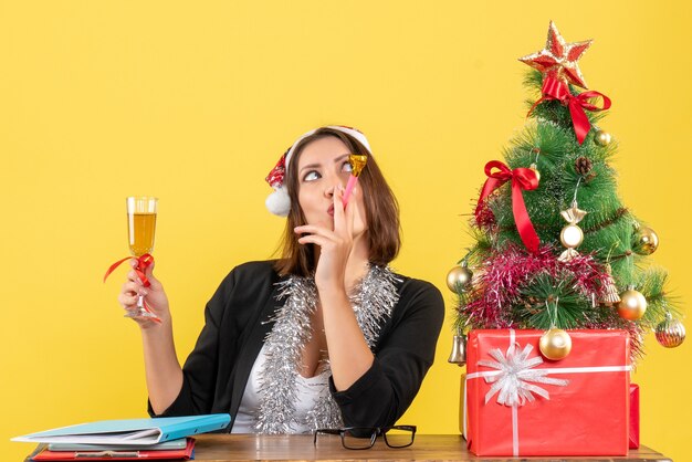 Emotionale glückliche charmante Dame im Anzug mit Weihnachtsmannhut und Neujahrsdekorationen, die Wein im Büro auf gelbem lokalisiertem Wein erheben