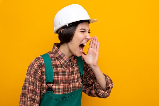 Emotionale junge Baumeisterin in Bauuniform und Schutzhelm, die jemanden schreit oder anruft, der die Hand in der Nähe des Mundes hält, der auf Orange steht