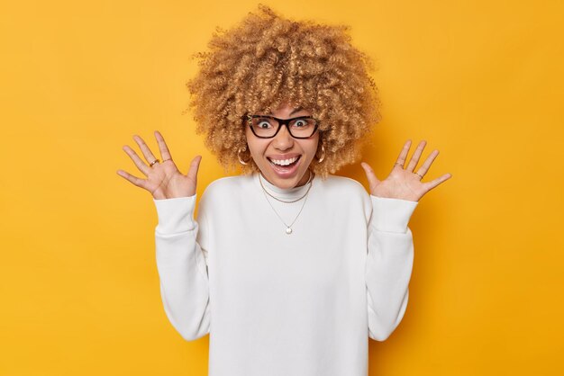 Emotionale junge Europäerin mit lockigen Haaren hält die Handflächen hoch, fühlt sich sehr glücklich, lächelt zahnlos und erhält tolle Neuigkeiten, trägt einen weißen Pullover und eine Brille, die auf leuchtend gelbem Hintergrund isoliert sind