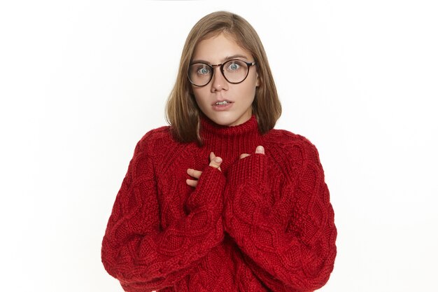 Emotionale junge Frau mit Käferaugen, die einen langärmeligen Strickpullover und eine Brille trägt, die einen überraschten Blick hat, den Mund öffnet und unerwartete Neuigkeiten erhält. Menschliche Mimik, Emotionen und Gefühle