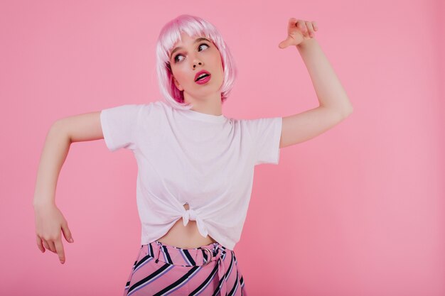 Emotionale junge Frau trägt kurze rosa Perücke herumalbern. Frohes weißes Mädchen in der Perücke lustiges Tanzen auf heller Wand