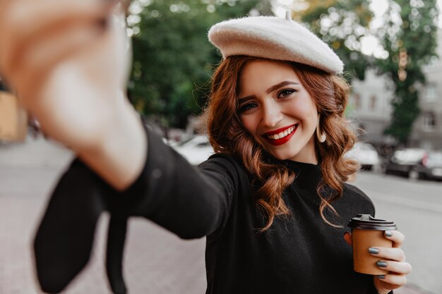 Emotionale kaukasische Frau, die selfie macht, während Tee im Herbst trinkt. Frohes Ingwermädchen trägt Baskenmütze, die Oktobertag genießt.