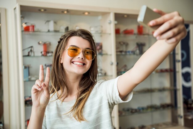 Emotionale modische urbane Frau im Optikerladen, der über Ständen mit Brille steht, während Selfie in stilvoller Sonnenbrille nimmt