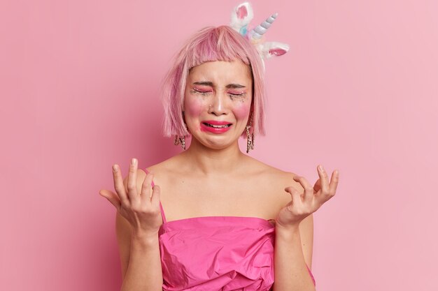 Emotionaler Burnout. Depressive gestresste rosahaarige Frau weint und sieht elend aus hebt die Hände aus Verzweiflung hat Make-up durchgesickert trägt Einhorn-Stirnbandkleid