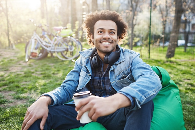 Emotionaler dunkelhäutiger Mann mit Afro-Frisur, der auf einem Stuhl im Park sitzt, mit jemandem spricht und Kaffee trinkt, breit lächelt und positive Emotionen ausdrückt.